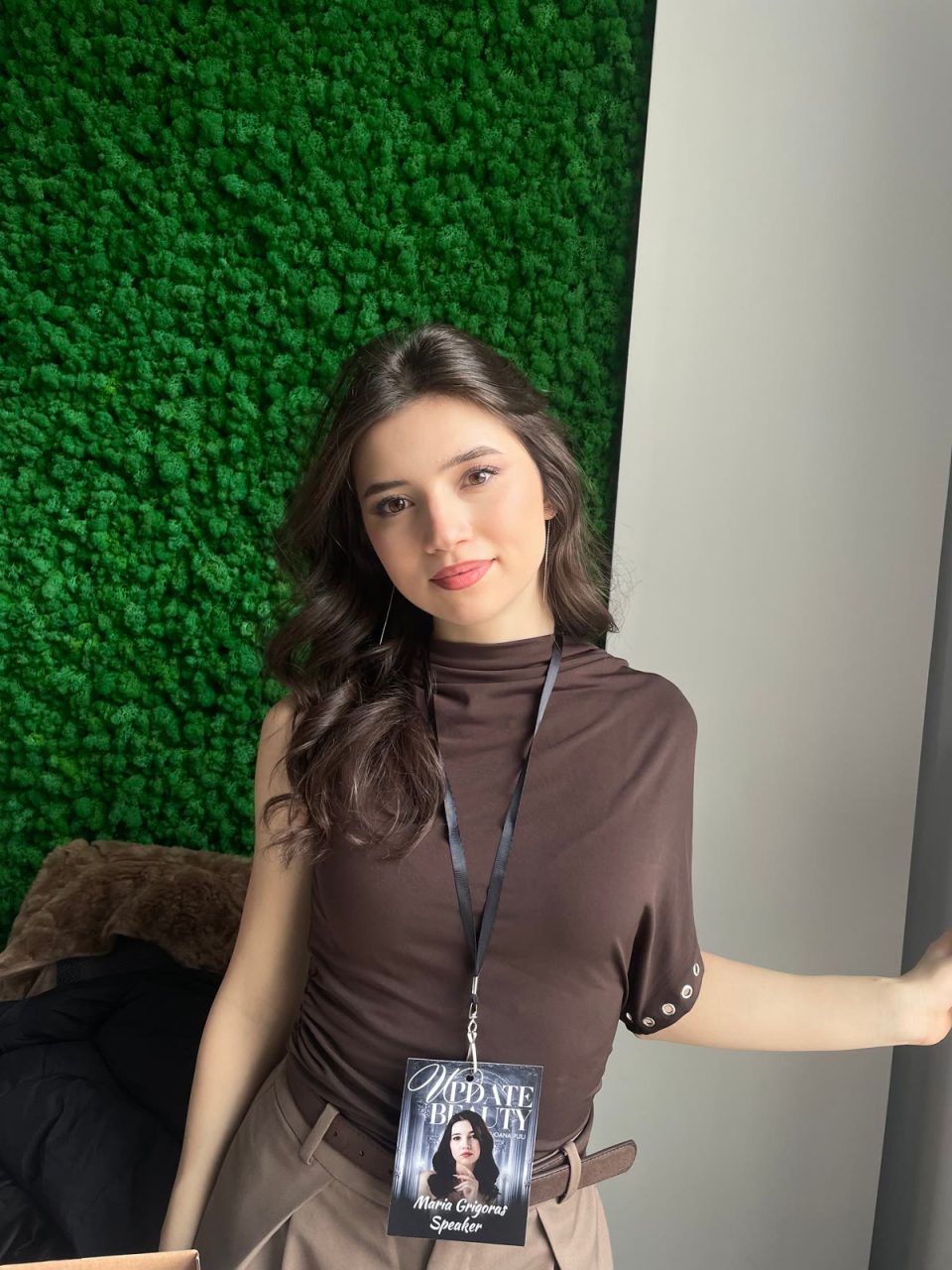 Maria Grigoraș zâmbind, în bluză maro, în fața unui perete de mușchi verde, cu ecusonul de speaker la gât